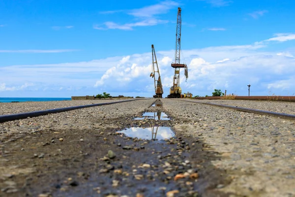view from a low point on the site of an old abandoned pier, in the foreground a puddle reflects a crane in it. broken shipping line