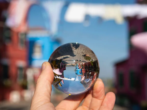 Hand holding glass sphere in front of colorful houses in Burano, Venice (Italy), sunny day in late autumn