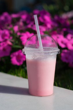Vertical photo of a pink milkshake in a plastic cup with a straw against the background of flowers in strong defocus
