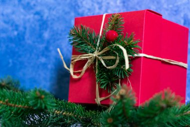 Kırmızı renkli bir hediye kutusunun yakın plan fotoğrafı kozalaklı dalların üzerinde duruyor, soyut mavi desenli bir duvara karşı..