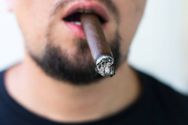 Sakallı bir adam dişlerinde yanan bir puro tutar. Arka plandaki adamın yüzü tamamen bulanık..