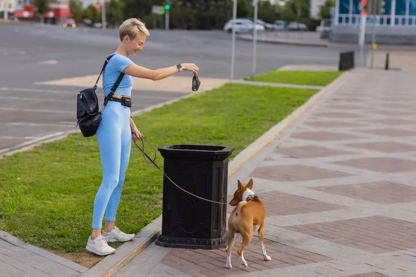 People working as dog-sitter, girl with french poodle dog in park. The young hispanic woman picks up her pets poo with plastic bag.