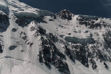 Karla kaplı dağ duvarının minimalist manzarası kalın alçak bulutların altında gün ışığında. Mavi gökyüzündeki yoğun bulutların arasında beyaz-kar zirvesi olan manzaralı parlak dağ manzarası. Karlı üstüyle harika bir manzara.