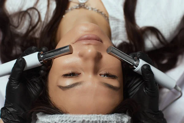 Kosmetologia. Piękna kobieta w klinice uzdrowiskowej otrzymująca od terapeuty stymulację elektrycznego leczenia twarzy. — Zdjęcie stockowe