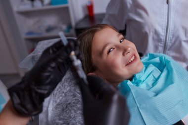 Diş hekimindeki küçük kız diş etlerine lokal anestezi yaptırıyor. Diş etlerini uyuşturan diş hekiminin kırpılmış görüntüsü. Pediatrik diş sağlığı konsepti.