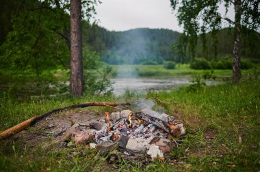 Gökyüzünün altındaki nehir kıyısında şenlik ateşi. Pembe bulutlar suya yansıyor, akşam alacakaranlığı..