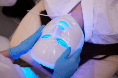 Yavaş çekimde yüz bakımı için hafif yaşlanma önleyici maske takıyordu. Özel maskeli bir kadın kanepede yatıyor. Modern güzellik ve sağlık teknolojileri.