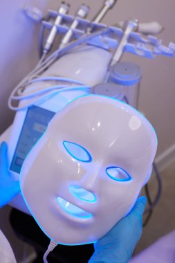 Yavaş çekimde yüz bakımı için hafif yaşlanma önleyici maske takıyordu. Özel maskeli bir kadın kanepede yatıyor. Modern güzellik ve sağlık teknolojileri.