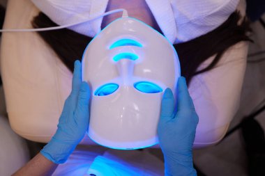 Yavaş çekimde yüz bakımı için hafif yaşlanma önleyici maske takıyordu. Özel maskeli bir kadın kanepede yatıyor. Modern güzellik ve sağlık teknolojileri.