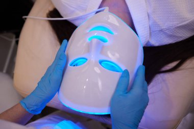 Yavaş çekimde yüz bakımı için hafif yaşlanma önleyici maske takıyordu. Özel maskeli bir kadın kanepede yatıyor. Modern güzellik ve sağlık teknolojileri.