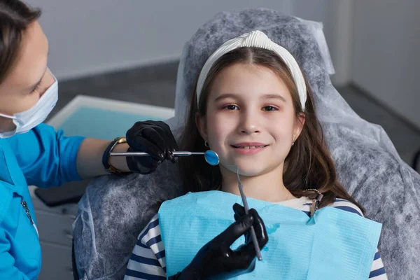Il dentista esamina i denti da latte delle neonate, il trattamento dei denti da latte, il dentista tiene uno specchio nelle sue mani, la carie in un bambino. — Foto Stock