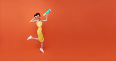 Zıplayan genç bir kadın su tabancasıyla ve Songkran festivali sırasında fotokopi çekilen turuncu arka planda mutlu bir güzellik..