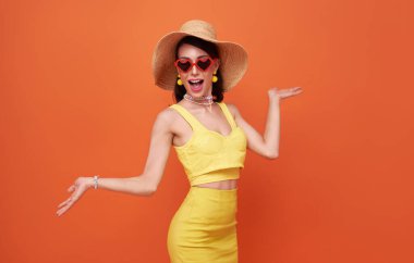 Happy attractive woman in summer dress and hat posing while standing and looking at camera isolated over orange background.