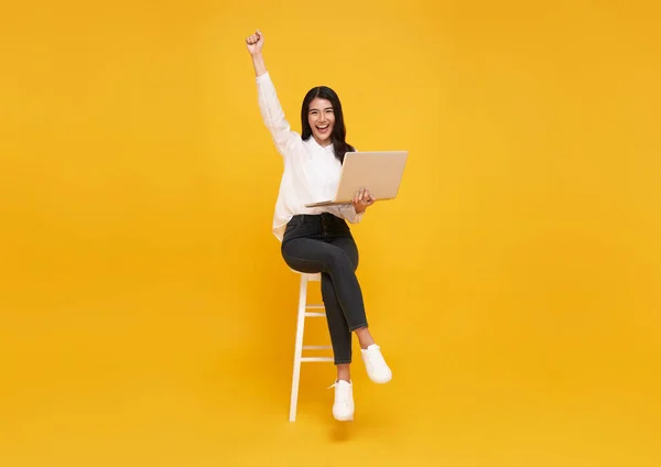 Young woman asian happy smiling celebrate. While her using laptop sitting on white chair isolate on bright yellow background.
