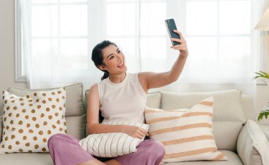 Evdeki kanepede rahat gülümseyen Asyalı kadın video görüşmesi için akıllı telefon kullanıyor..