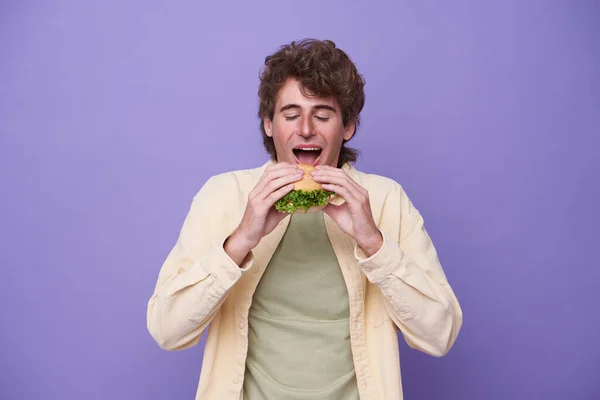 拿着一块汉堡包的年轻人在紫色背景下吃孤立的东西 — 图库照片