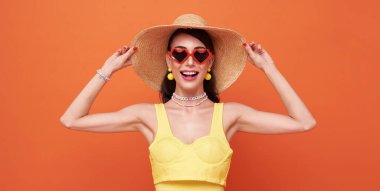 Happy attractive woman in summer dress and hat posing while standing and looking at camera isolated over orange background.