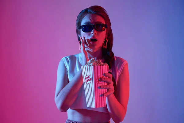Young woman wearing 3D glasses holding popcorn and beverage watching exciting movie isolated on neon background.