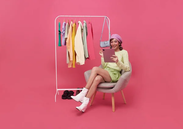 若い笑顔の美しいアジアの女性がオンラインショッピングを作り ピンクの背景に孤立した新しい服で椅子に座っています オンラインショッピング — ストック写真