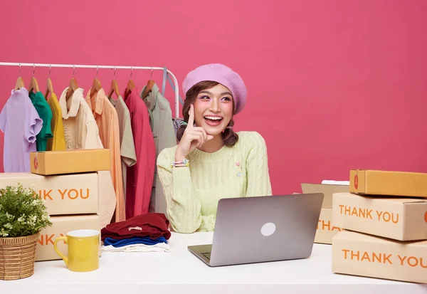 Young asian woman startup small business freelance sale fashion clothing with parcel box and computer laptop on table and sitting isolated on pink background. Online marketing and delivery concept.