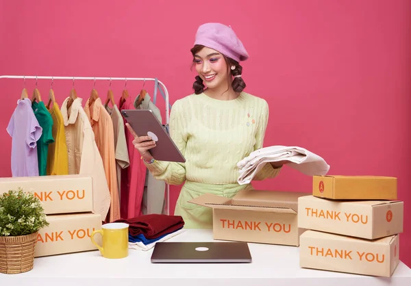 Young asian woman startup small business freelance sale fashion clothing with parcel box and computer laptop on table and sitting isolated on pink background. Online marketing and delivery concept.