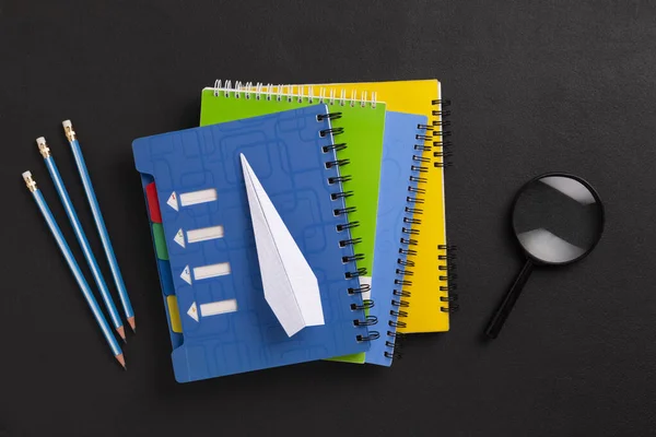 The concept of education and future planning. Close-up of a stack of colored notebooks, a magnifying glass, pencils and a paper airplane - a symbol of dreams