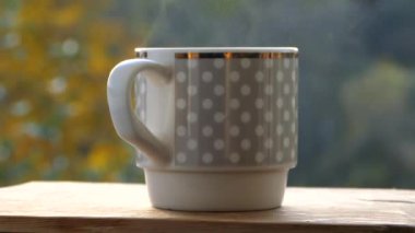 Sonbahar havası ve sarı ağaçların arka planında fincandan buhar yükselir. Sıcak sonbaharda dışarıda sıcak çay iç.