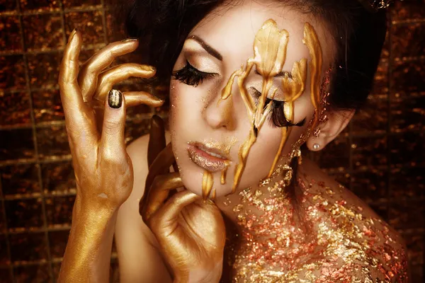 Retrato de close-up de bela jovem morena com pestanas de penas longas em tinta líquida dourada e folha de bronze no fundo dourado segurando as mãos perto de seu rosto — Fotografia de Stock