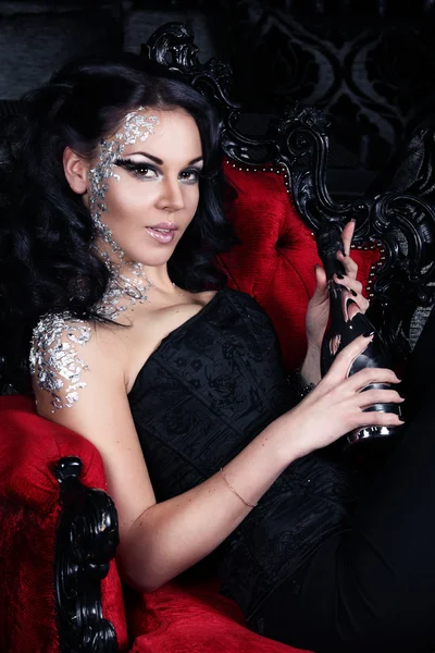 baştan çıkarıcı esmer güzel kırmızı Gotik sandalyede oturan ve gümüş bir şişe şampanya holding.
