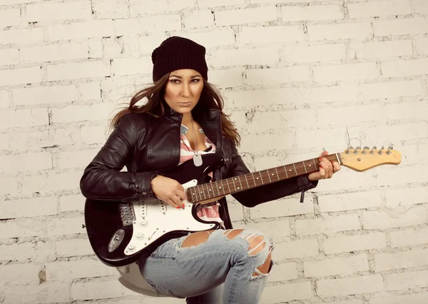 Jovem adolescente procurando mulher tocando sua guitarra eletrônica preta usando sua jaqueta de couro e gorro de malha de pé na frente da parede de tijolo branco — Fotografia de Stock