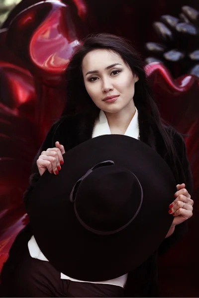 Attractive asian young female is holding her black hat in her hands on the red background and looking to the camera — Stock Photo, Image