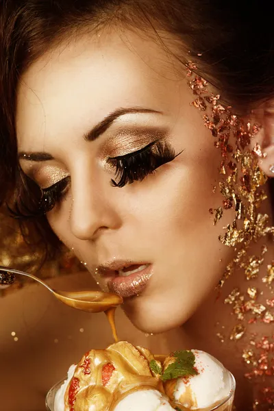 Portrait of beautiful brunette with long lashes and golden makeup is holding a spoon and eating caramel ice cream with strawberry and mint — Stock Photo, Image