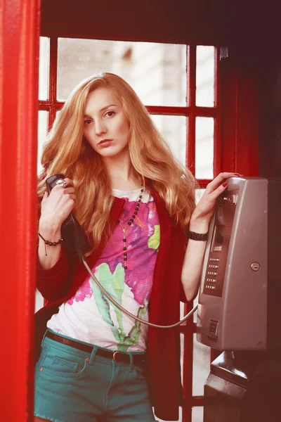 Belle femme britannique aux cheveux roux clair se tient debout dans la cabine téléphonique traditionnelle de Londres avec le téléphone dans ses mains et regardant vers l'appareil photo — Photo