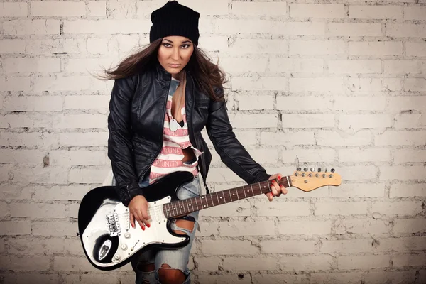 Young teenage looking woman playing her black electronic guitar wearing her leather jacket and knitted beanie standing in front of white brick wall — Stock Photo, Image