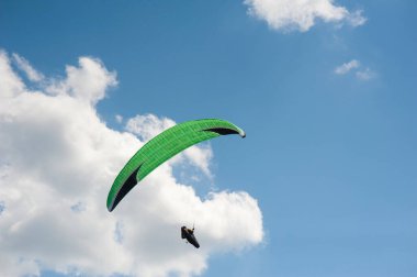 Bulutların arka planında mavi gökyüzünde uçan tek başına yeşil bir paraglider. Güneşli bir günde gökyüzünde paraşütle atlamak.