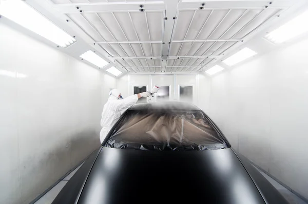 Trabajador pintando un coche negro en un garaje especial, con un traje blanco y equipo de protección — Foto de Stock