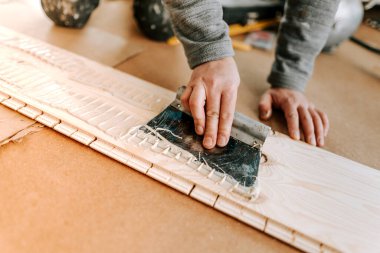 close up of worker hands, craftsman using glue and trowel for spreading glue on wooden board of hardwood parquet