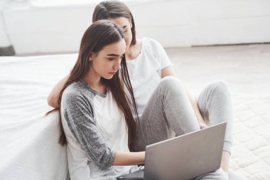 Rahatlatıcı ve rahatlatıcı. Genç bayan ikizler beyaz yatağın yanında oturuyor ve gümüş renkli dizüstü bilgisayar kullanıyorlar..