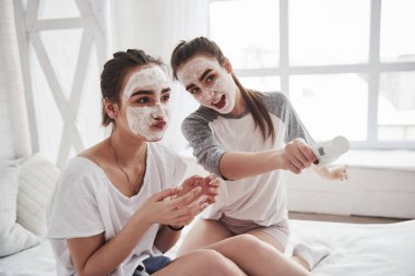 Neşeli bir ruh hali. Yüzünde beyaz maske kullanarak cilt bakımı kavramı. İki kız kardeş hafta sonunu yatak odasında geçirir..