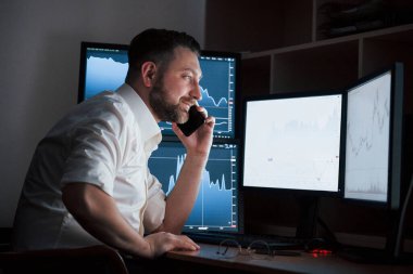 Keyif alıyorum. Beyaz gömlekli sakallı adam ofiste çalışıyor. İndeks çizelgelerinde birden fazla bilgisayar ekranı var..