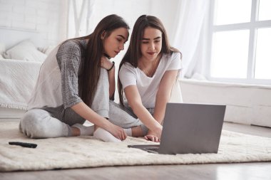 Genç bayan ikizler beyaz yatağın yanında oturuyor ve gümüş renkli dizüstü bilgisayar kullanıyorlar..