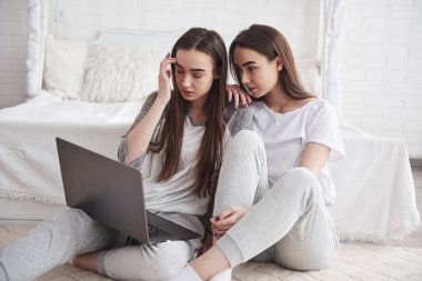 Ön manzara. Genç bayan ikizler beyaz yatağın yanında oturuyor ve gümüş renkli dizüstü bilgisayar kullanıyorlar..
