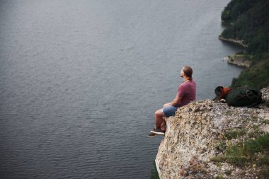 Sırt çantalı gezgin dağın tepesinde oturmuş su yüzeyinin keyfini çıkarıyor. Dağlar ve kıyı boyunca seyahat, özgürlük ve aktif yaşam tarzı konsepti.