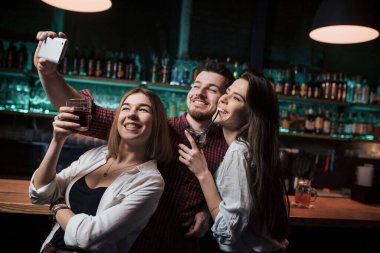 Arkasında alkol olan bir sürü şişe. Arkadaşlar güzel bir gece kulübünde selfie çekiyorlar. İçkiler ellerde.