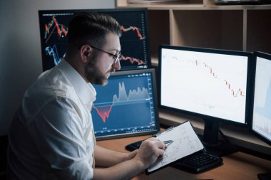 Not defteri kullanıyor. Beyaz gömlekli sakallı adam ofiste çalışıyor. İndeks çizelgelerinde birden fazla bilgisayar ekranı var..