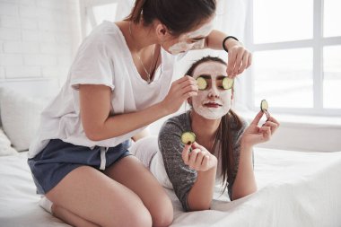 Yüzüne salatalık sürüyorum. Beyaz maske kullanarak cilt bakımı kavramı. İki kız kardeş hafta sonunu yatak odasında geçirir..