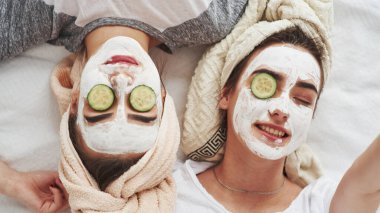 Gözümü açamıyorum. Taze salatalık halkaları ve yüzünde beyaz maske kullanarak cilt bakımı kavramı. İki kız kardeş hafta sonunu yatak odasında geçirir..