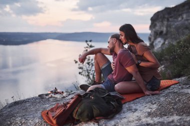 Sırt çantalı çift dağın tepesinde oturmuş nehir veya göl manzarasının keyfini çıkarıyorlar. Dağlar ve kıyı boyunca seyahat, özgürlük ve aktif yaşam tarzı konsepti.