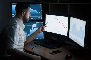 Bilgiyi göstermek için kalem kullanıyor. Beyaz gömlekli sakallı adam ofiste çalışıyor. İndeks çizelgelerinde birden fazla bilgisayar ekranı var..