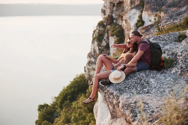 岩の山の上に座って川や湖の海岸の景色を楽しむバックパック付きの巨大なカップル 山や海岸を旅し 自由とアクティブなライフスタイルのコンセプト — ストック写真